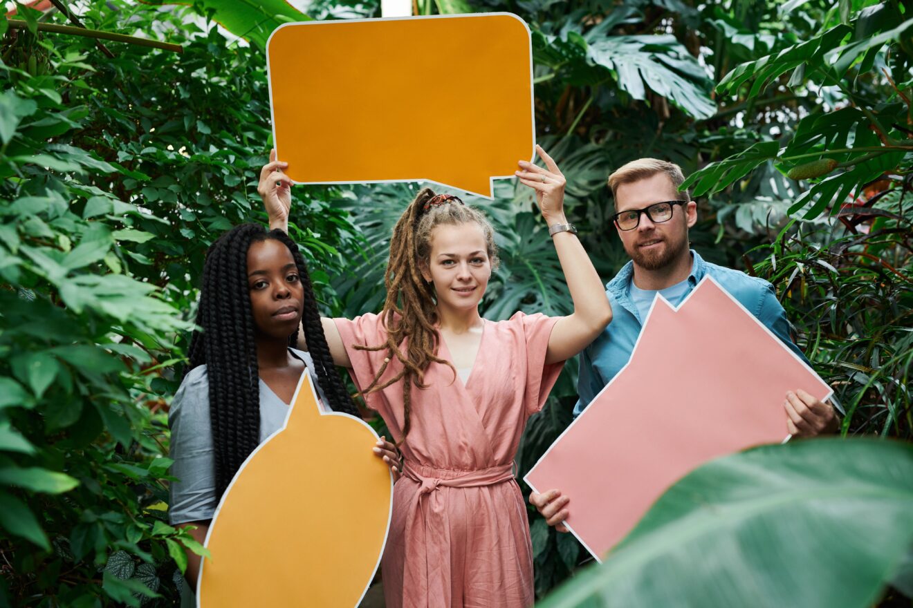 Three adults of diverse backgrounds holding colorful speech bubbles amidst lush greenery.