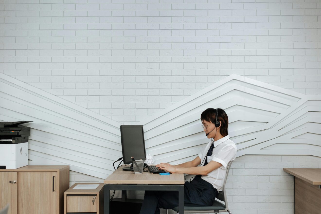 Asian woman in office wearing headset, focused on computer tasks at her desk.